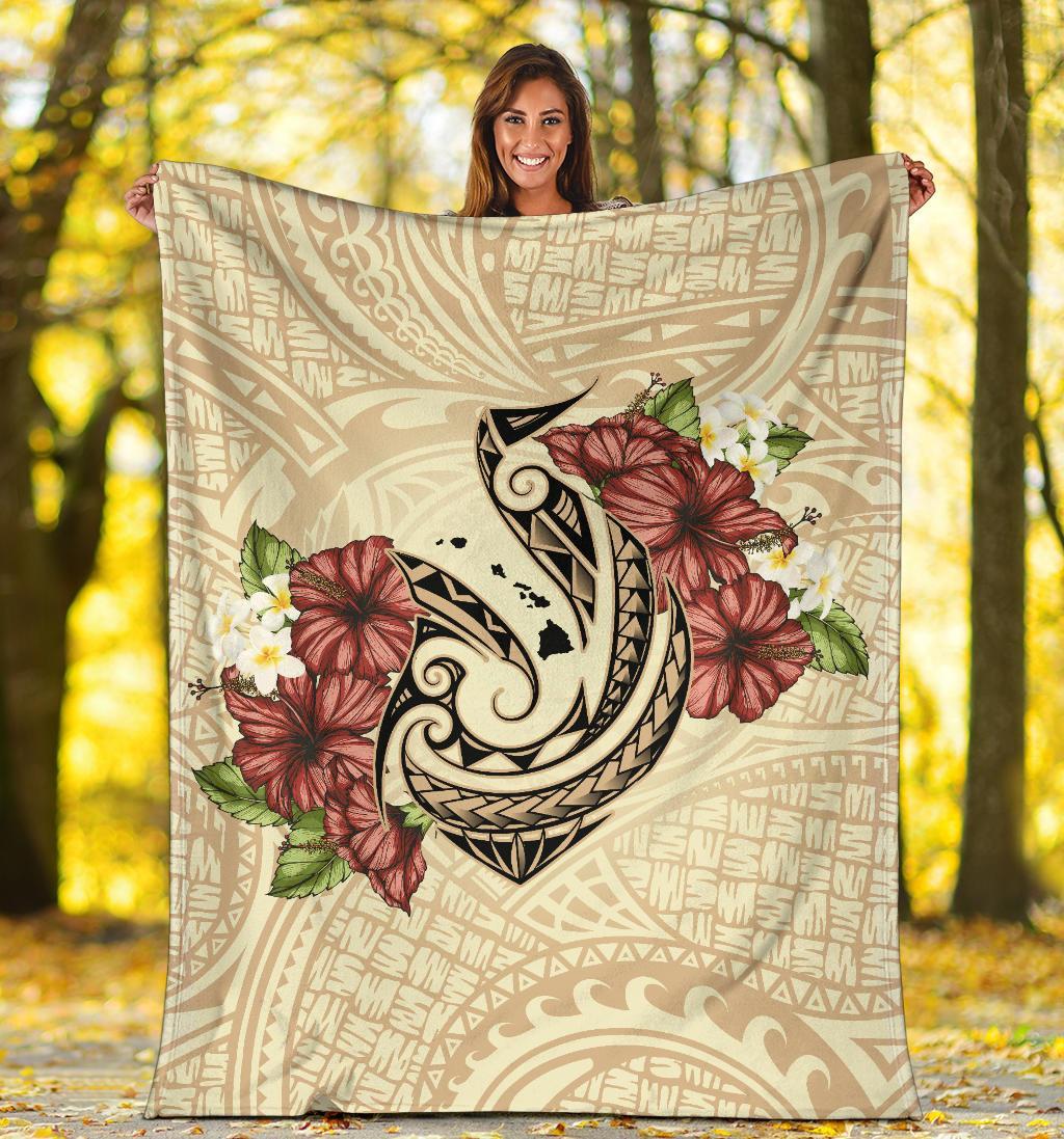 Beige blanket held outdoors with fish hook emblem, red hibiscus blooms, and white flowers