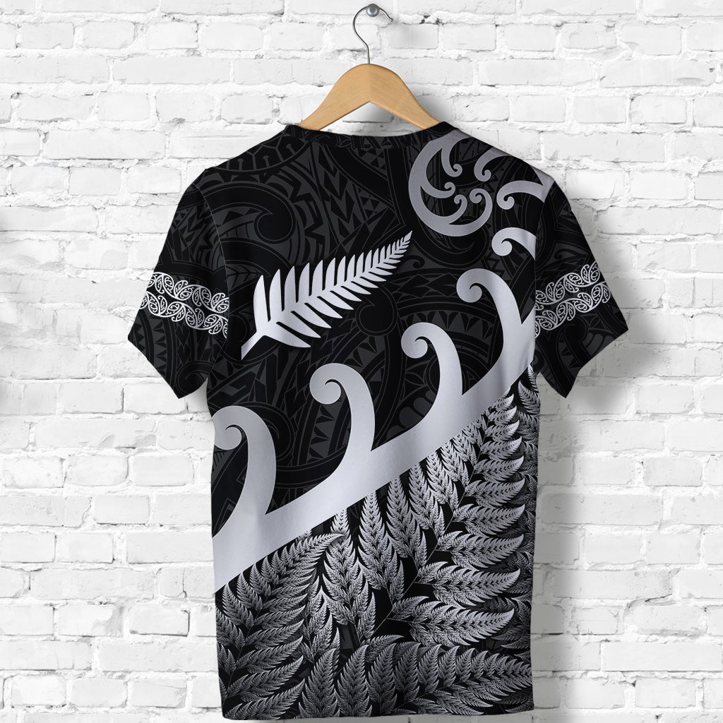 Black T-shirt with white koru swirls and fern-leaf band over textured leaves