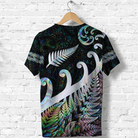 Dark green T-shirt with white fern-and-koru design across front, colorful feather pattern